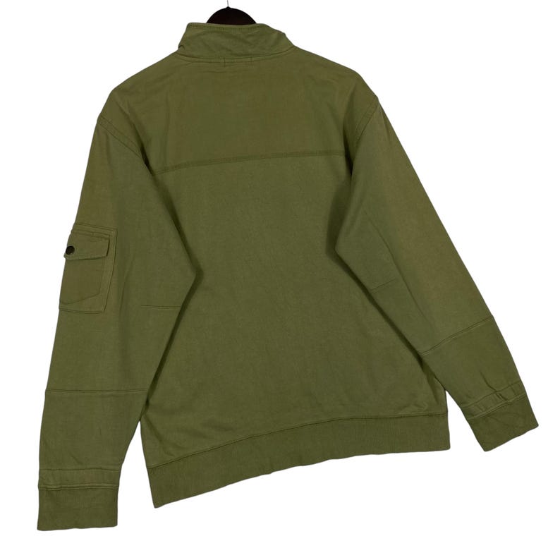 Puede incluir: Sudadera con cremallera verde oliva con un peque&ntilde;o bolsillo en la manga izquierda. La sudadera tiene un cuello alto y dobladillos y pu&ntilde;os acanalados. Vista trasera que muestra las costuras y los detalles de construcci&oacute;n.