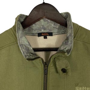 Puede incluir: Chaqueta verde oliva con cremallera y cuello alzado. La chaqueta presenta un estampado de camuflaje en el cuello y el logotipo de Van Dutch. La chaqueta est&aacute; colgada en una percha de madera.