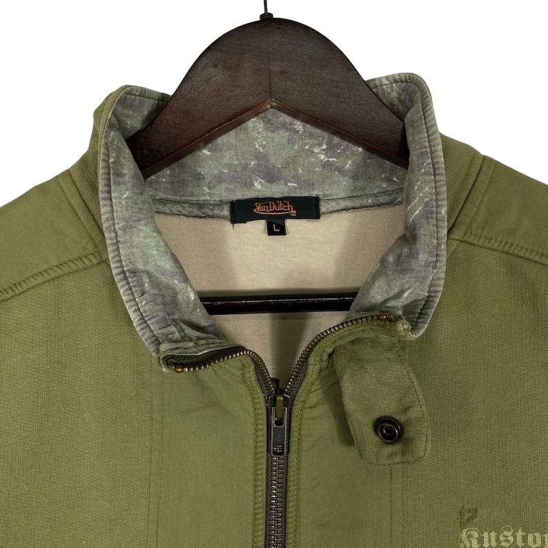 Puede incluir: Chaqueta verde oliva con cremallera y cuello alzado. La chaqueta presenta un estampado de camuflaje en el cuello y el logotipo de Van Dutch. La chaqueta est&aacute; colgada en una percha de madera.
