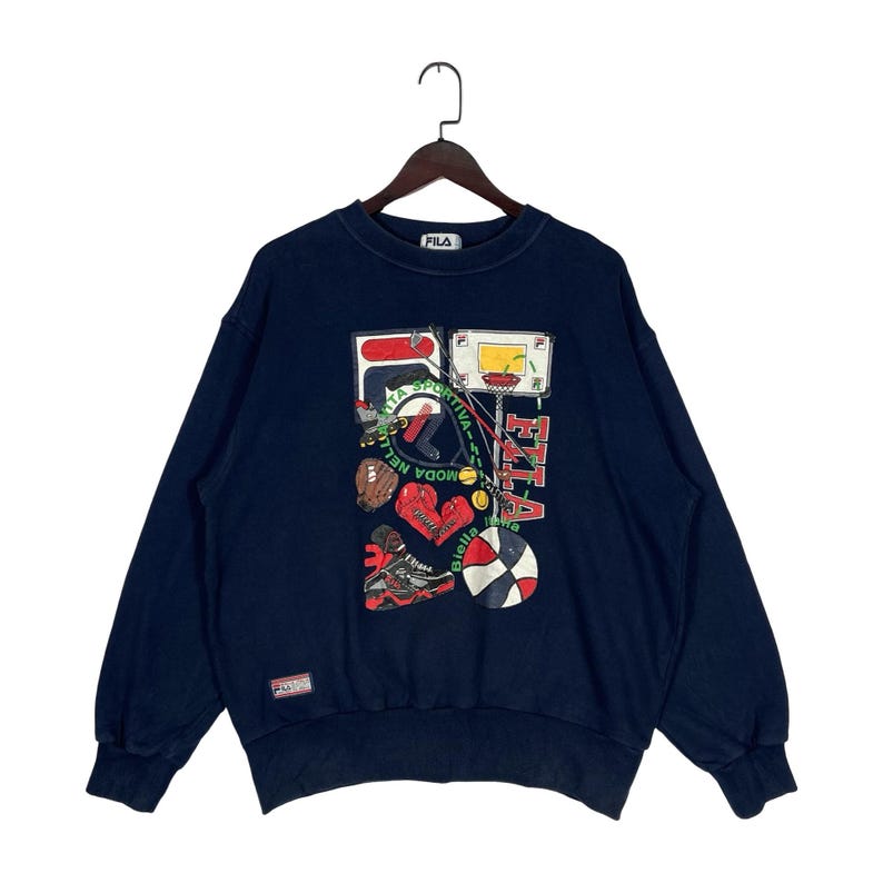 May include: A navy blue long-sleeve sweatshirt featuring a graphic print of various sports equipment including a basketball hoop, boxing gloves, a tennis racket, and a sneaker. The Fila logo is printed in red and white, with additional text.