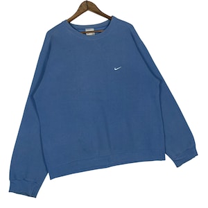 以下が含まれることがあります： 胸に小さな白いナイキのスウッシュロゴが入った青いクルーネックのスウェットシャツ。スウェットシャツは木製のハンガーに吊るされています。生地は柔らかく快適な素材のようです。