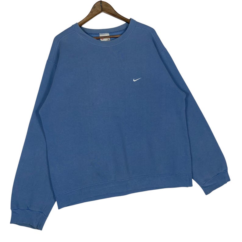 以下が含まれることがあります： 胸に小さな白いナイキのスウッシュロゴが入った青いクルーネックのスウェットシャツ。スウェットシャツは木製のハンガーに吊るされています。生地は柔らかく快適な素材のようです。