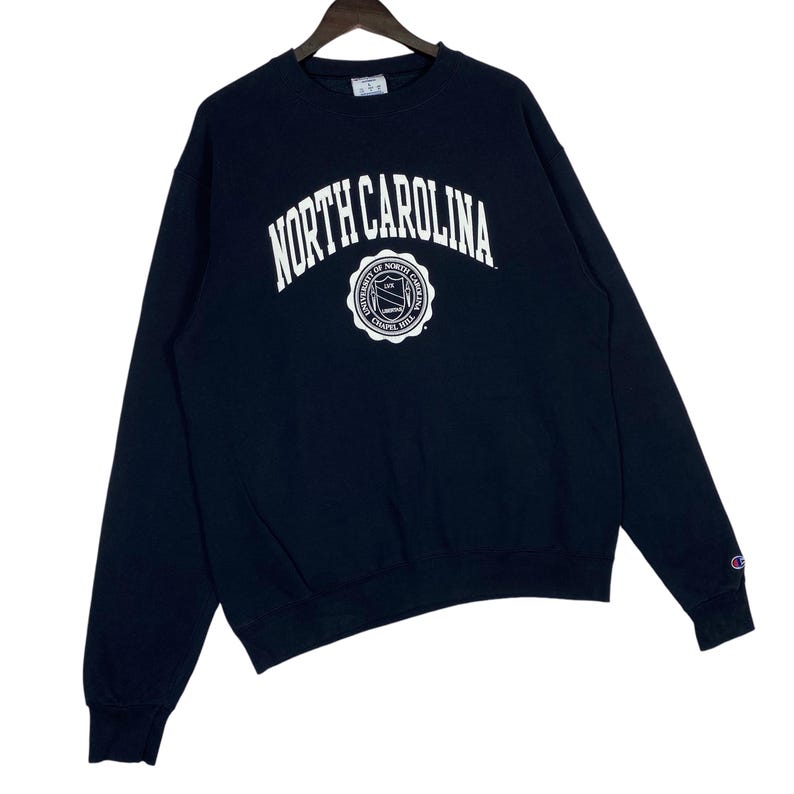 May include: Navy blue Champion sweatshirt featuring the words "NORTH CAROLINA" in white arched lettering above a circular emblem. The emblem includes text and a seal design. The sweatshirt has long sleeves and a crew neck.