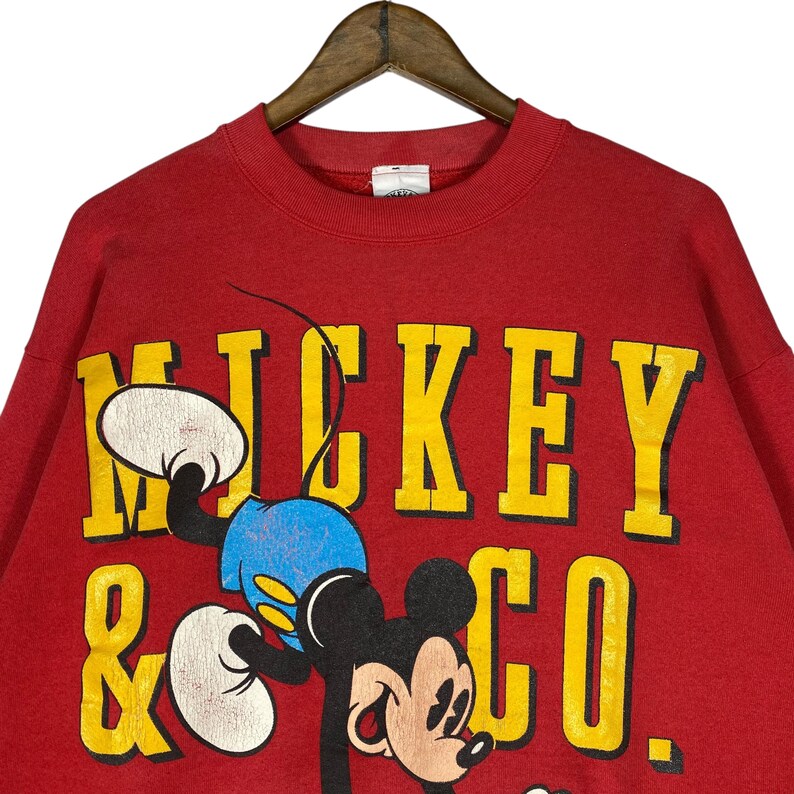 May include: Red sweatshirt featuring a large yellow graphic that reads "MICKEY & CO." with a cartoon image of Mickey Mouse. The sweatshirt has a crew neck and long sleeves. The graphic includes Mickey Mouse in blue shorts and white gloves.