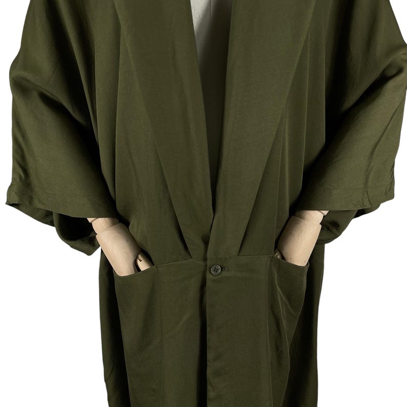 Puede incluir: Bata estilo kimono verde oliva con mangas anchas y cuello chal. La bata tiene dos bolsillos delanteros y un cierre de bot&oacute;n &uacute;nico en la cintura. La tela tiene un brillo sutil, lo que sugiere un material ligero.