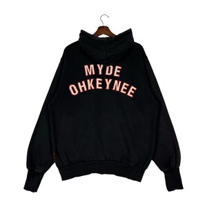 May include: Black hoodie with the text "MYDE OHKEYNEE" in red and white lettering across the back. The hoodie has a hood, long sleeves, and ribbed cuffs. The garment is hanging on a wooden hanger.