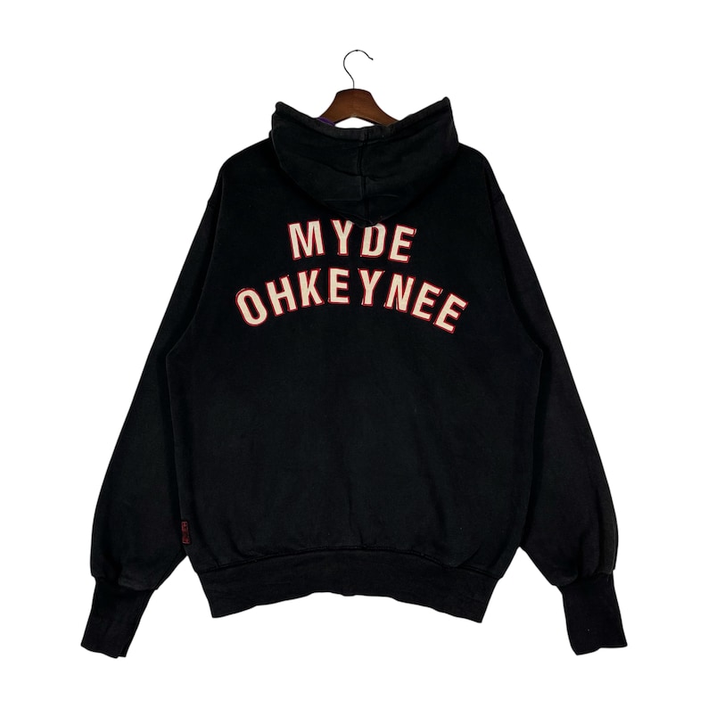May include: Black hoodie with the text "MYDE OHKEYNEE" in red and white lettering across the back. The hoodie has a hood, long sleeves, and ribbed cuffs. The garment is hanging on a wooden hanger.