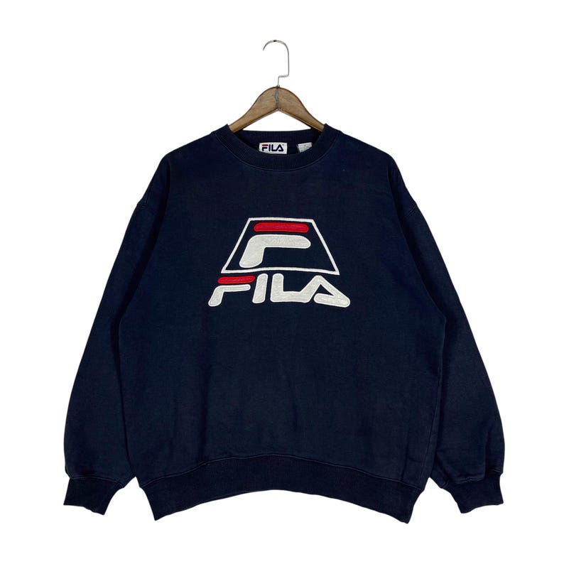 Puede incluir: Sudadera azul marino con el logotipo de FILA en blanco, rojo y negro. El logotipo est&aacute; centrado en el pecho y presenta el nombre de la marca en una fuente estilizada. La sudadera tiene cuello redondo y mangas largas.