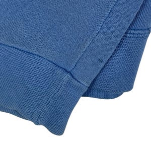 以下が含まれることがあります： 明るい青色のニット生地のクローズアップ。おそらく衣服の袖口または袖口。生地は袖口にリブ編みのテクスチャがあり、体に滑らかでプレーンなニットがあります。画像はステッチの詳細と生地の色を示しています。