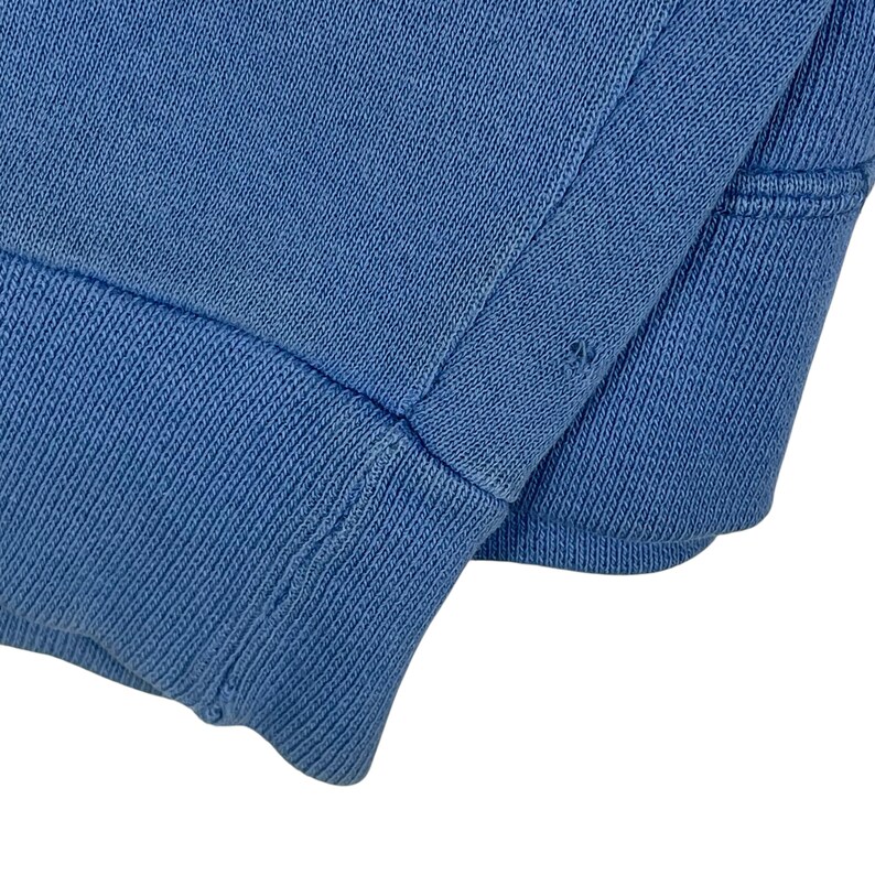 以下が含まれることがあります： 明るい青色のニット生地のクローズアップ。おそらく衣服の袖口または袖口。生地は袖口にリブ編みのテクスチャがあり、体に滑らかでプレーンなニットがあります。画像はステッチの詳細と生地の色を示しています。