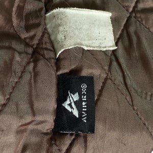 May include: Close-up of brown quilted fabric with a white tag and a black label. The black label displays the brand name "AVIREX&reg;" and a stylized logo. The fabric has a diamond pattern.