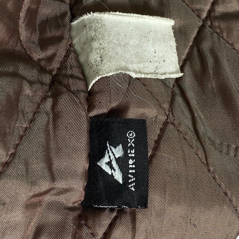 May include: Close-up of brown quilted fabric with a white tag and a black label. The black label displays the brand name "AVIREX&reg;" and a stylized logo. The fabric has a diamond pattern.