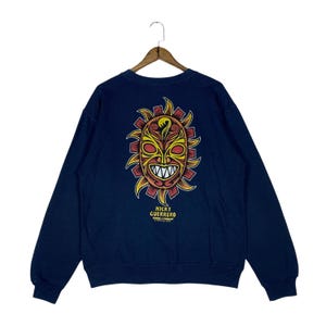 May include: Navy blue sweatshirt featuring a sun-faced mask design in yellow, red, and orange. The text "NICKY GUERRERO" is printed below the graphic. The sweatshirt is suspended from a wooden hanger.
