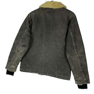 May include: A grey denim jacket featuring a tan sherpa collar. The jacket has black cuffs and a worn look. The back view of the jacket is shown, highlighting the stitching and construction details. A classic outerwear piece.