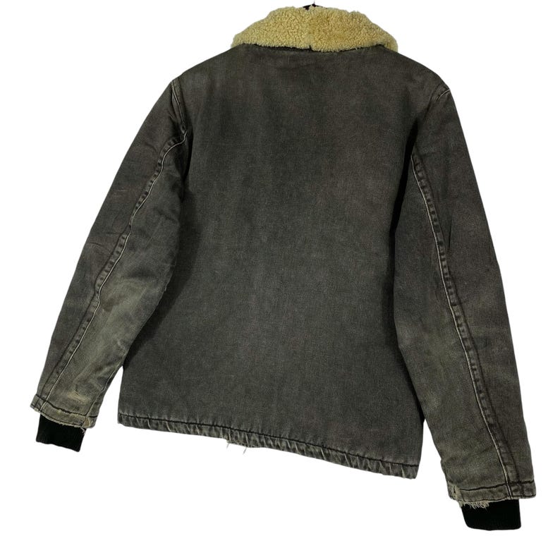 May include: A grey denim jacket featuring a tan sherpa collar. The jacket has black cuffs and a worn look. The back view of the jacket is shown, highlighting the stitching and construction details. A classic outerwear piece.