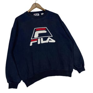 Puede incluir: Sudadera azul marino con el logo de FILA en blanco, rojo y negro. El logo es un dise&ntilde;o geom&eacute;trico estilizado. La sudadera tiene cuello redondo y mangas largas. El logo de FILA est&aacute; centrado en el pecho.