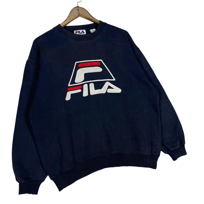 Puede incluir: Sudadera azul marino con el logo de FILA en blanco, rojo y negro. El logo es un dise&ntilde;o geom&eacute;trico estilizado. La sudadera tiene cuello redondo y mangas largas. El logo de FILA est&aacute; centrado en el pecho.