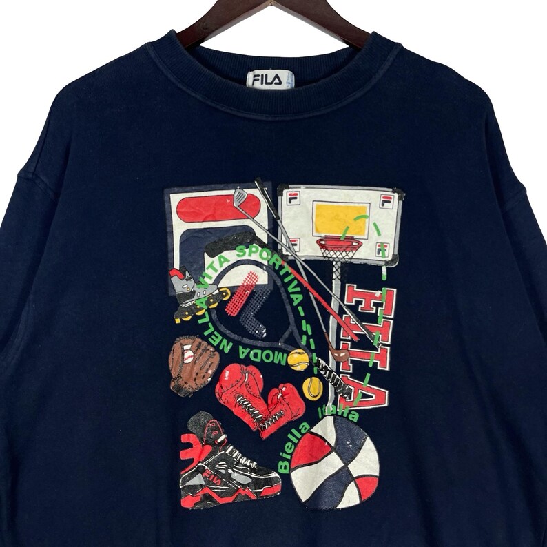 May include: A navy blue crewneck sweatshirt featuring a large graphic print on the front. The design includes various sports equipment such as a basketball hoop, boxing gloves, a baseball glove, a tennis racket, and a sneaker, with the text FILA.