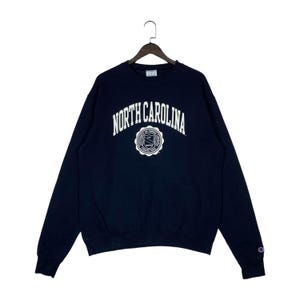 May include: Navy blue Champion sweatshirt with the words "NORTH CAROLINA" in white arched lettering. Below the text is a white circular emblem with text and a shield. The sweatshirt has long sleeves and a crew neck.