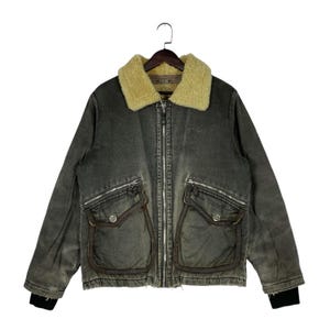 May include: A distressed grey jacket with a tan shearling collar, two front pockets, and a zip closure. The jacket has a vintage look and black ribbed cuffs. The jacket is hanging on a wooden hanger.