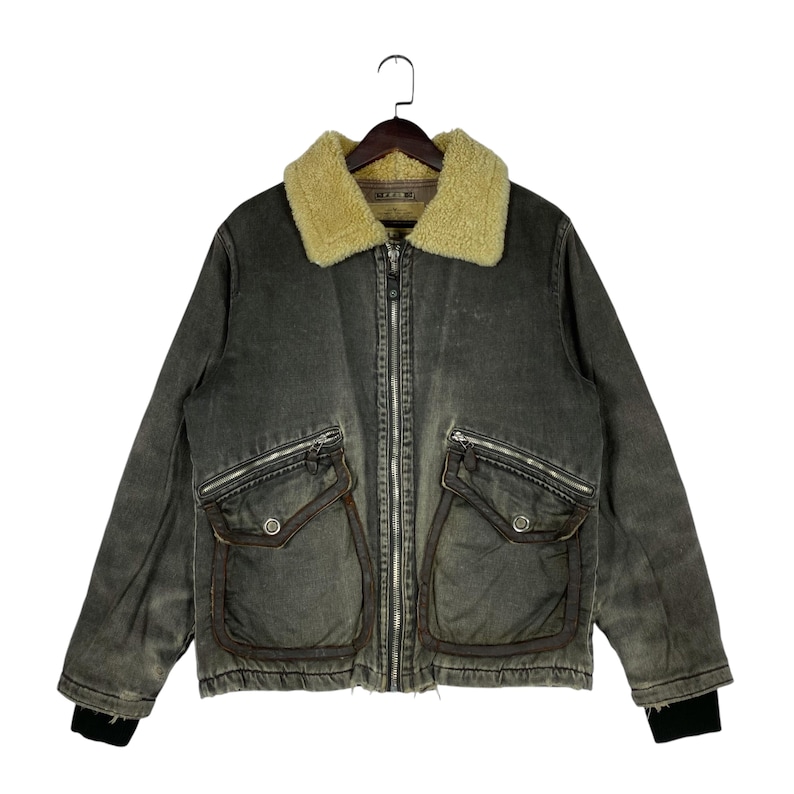 May include: A distressed grey jacket with a tan shearling collar, two front pockets, and a zip closure. The jacket has a vintage look and black ribbed cuffs. The jacket is hanging on a wooden hanger.