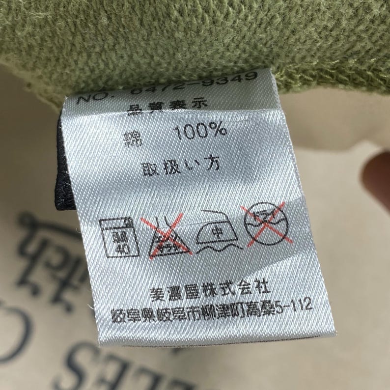 Puede incluir: Primer plano de una etiqueta de tela blanca con texto japon&eacute;s y s&iacute;mbolos de cuidado. La etiqueta indica 100% algod&oacute;n e instrucciones de lavado. La etiqueta est&aacute; adherida a una tela verde.