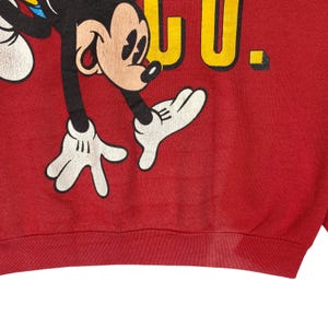 May include: Red sweatshirt featuring a graphic of Mickey Mouse in black, white, and yellow. The word "CLUB" is printed in yellow. The sweatshirt has a ribbed hem and cuffs. The design is vintage and shows signs of wear.