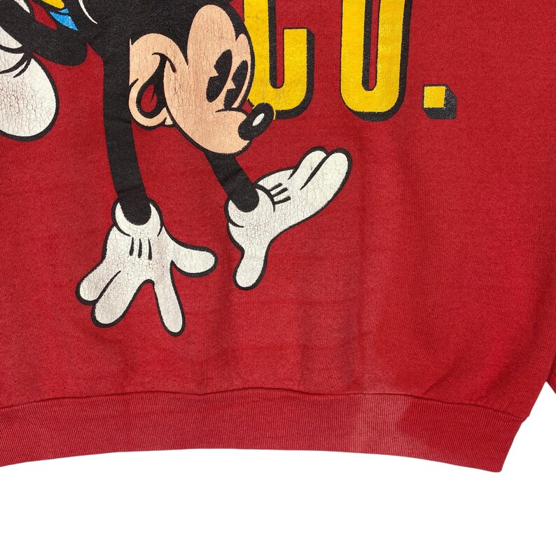 May include: Red sweatshirt featuring a graphic of Mickey Mouse in black, white, and yellow. The word "CLUB" is printed in yellow. The sweatshirt has a ribbed hem and cuffs. The design is vintage and shows signs of wear.