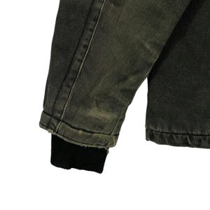 May include: Close-up of a distressed denim jacket sleeve. The jacket is a faded olive green colour with a black ribbed cuff. There is a small tear near the cuff, adding to the vintage aesthetic. The fabric shows signs of wear.