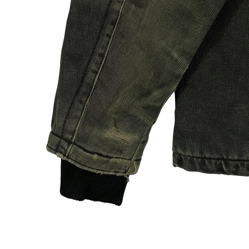 May include: Close-up of a distressed denim jacket sleeve. The jacket is a faded olive green colour with a black ribbed cuff. There is a small tear near the cuff, adding to the vintage aesthetic. The fabric shows signs of wear.