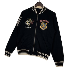 May include: Black zip-up jacket with white and beige striped trim. Features embroidered dragon and "AVIREX LTD" text on the front, and a U.S. Air Force emblem. The jacket has a full zipper and ribbed cuffs.