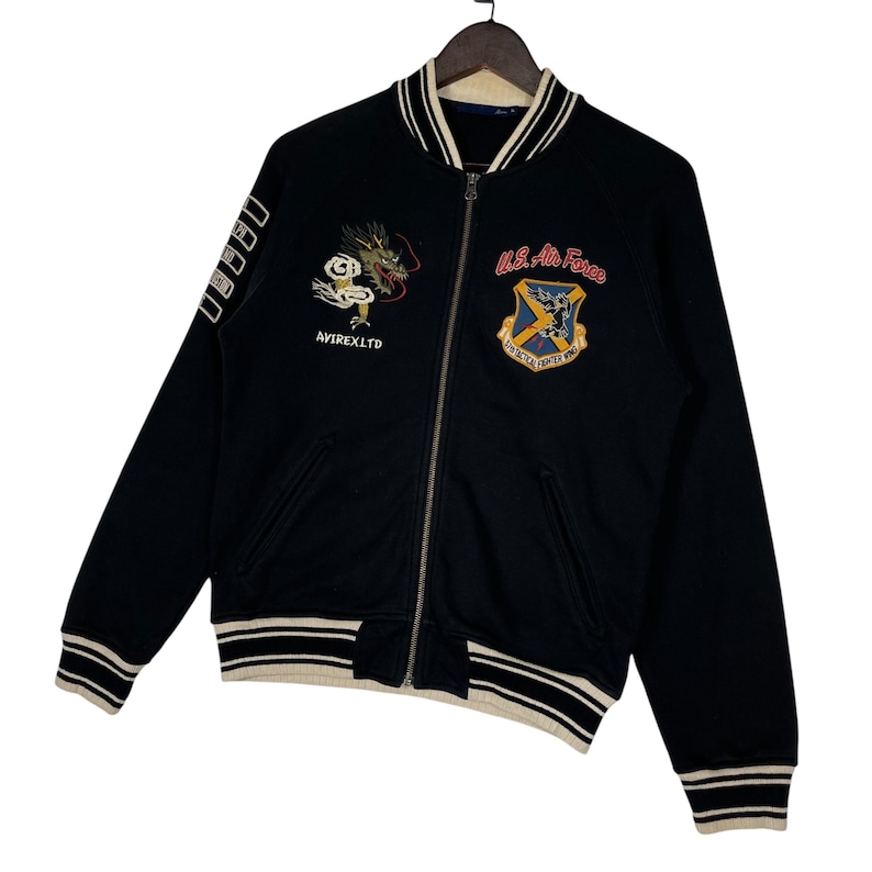 May include: Black zip-up jacket with white and beige striped trim. Features embroidered dragon and "AVIREX LTD" text on the front, and a U.S. Air Force emblem. The jacket has a full zipper and ribbed cuffs.