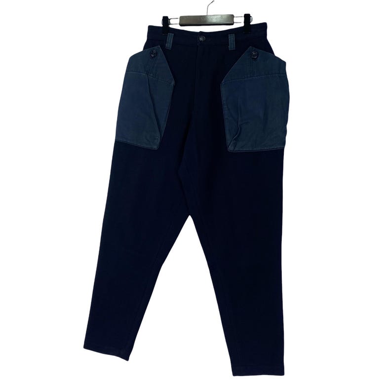 May include: Dark navy blue pants with two large, square, navy blue pockets on the back. The pants are hanging on a hanger against a white background. The pants have a button and zipper closure.