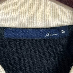 May include: Close-up of a black sweatshirt collar with a cream-coloured ribbed neckline. A navy blue label displays the brand name "A&auml;rex" in white script. The sweatshirt is likely made of a soft, comfortable material.