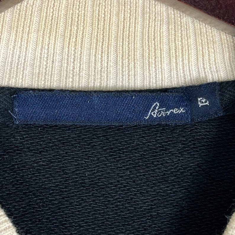 May include: Close-up of a black sweatshirt collar with a cream-coloured ribbed neckline. A navy blue label displays the brand name "A&auml;rex" in white script. The sweatshirt is likely made of a soft, comfortable material.