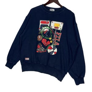 May include: A navy blue long-sleeve sweatshirt featuring a graphic print of various sports equipment including a basketball, boxing gloves, a baseball glove, and a basketball hoop. The text FILA and Biella Italia are printed on the front.