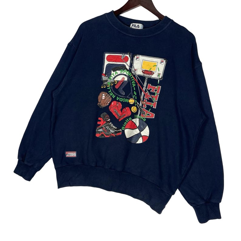 May include: A navy blue long-sleeve sweatshirt featuring a graphic print of various sports equipment including a basketball, boxing gloves, a baseball glove, and a basketball hoop. The text FILA and Biella Italia are printed on the front.
