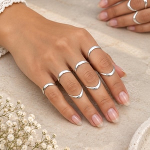 May include: A set of polished silver rings worn on multiple fingers. The rings feature a smooth, rounded band design with a subtle V-shaped curve on some pieces. The jewelry has a reflective metallic finish and is worn across both hands.