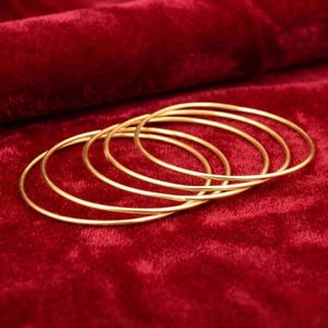 Może przedstawiać: Zestaw pięciu cienkich, okrągłych bransoletek typu bangle z metalu w kolorze złotym. Każda bransoletka ma gładką, polerowaną powierzchnię o jednolitym, okrągłym kształcie. Bransoletki są ułożone w nakładającą się na siebie, warstwową formację.