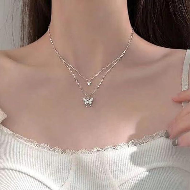 May include: A silver-toned, layered necklace with two butterfly pendants. The top layer features a small butterfly charm, and the lower layer has a larger, detailed butterfly. The necklace is designed to be worn around the neck.