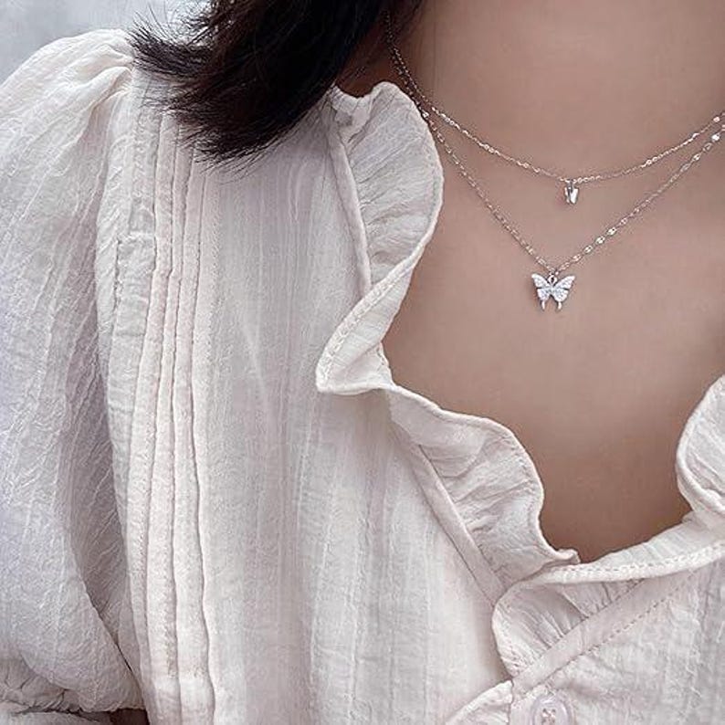 May include: A silver, layered necklace featuring butterfly pendants. The necklace is worn over a white blouse with frilled details. The butterfly pendants are small and delicate, adding a touch of elegance.