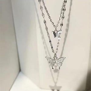 May include: A double-layered silver necklace with butterfly pendants. The lower pendant is a larger butterfly, embellished with small, sparkling stones. The necklace has a delicate chain design, suitable for adding elegance to any outfit.