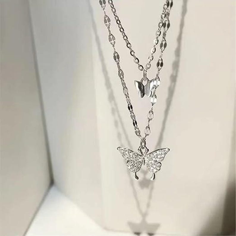 May include: A double-layered silver necklace with butterfly pendants. The lower pendant is a larger butterfly, embellished with small, sparkling stones. The necklace has a delicate chain design, suitable for adding elegance to any outfit.