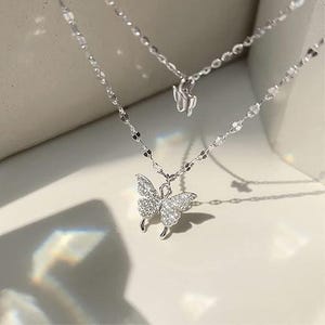 May include: A silver-coloured, layered necklace with butterfly pendants. The lower pendant is a butterfly covered in small, clear stones. The necklace has a delicate chain design, and the background is a neutral colour.