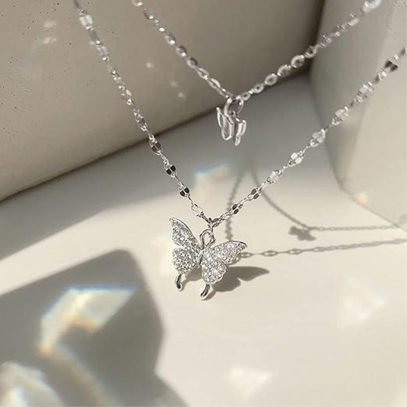 May include: A silver-coloured, layered necklace with butterfly pendants. The lower pendant is a butterfly covered in small, clear stones. The necklace has a delicate chain design, and the background is a neutral colour.