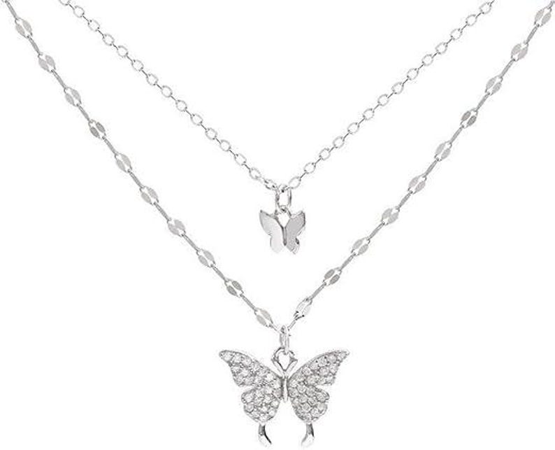 May include: A silver-coloured, layered necklace with two butterfly pendants. The top pendant is a small butterfly, and the lower pendant is a larger butterfly encrusted with small, clear stones. The necklace has two delicate chains.