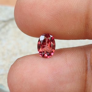 Puede incluir: Una piedra preciosa de corte ovalado con un color rojo intenso y una superficie facetada que refleja la luz. La piedra se sostiene entre dos dedos, mostrando su calidad transparente y forma simétrica. Las facetas crean un patrón geométrico.
