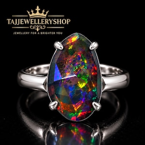 May include: A pear-shaped black opal ring set in a polished silver-toned metal band with four prongs. The opal displays vibrant flashes of red, green, and blue. The text TAJJEWELLERYSHOP JEWELLERY FOR A BRIGHTER YOU is visible at the top.