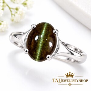 Puede incluir: Un anillo de plata con una piedra preciosa ovalada de color verde oscuro con efecto ojo de gato, montada en un engaste de cuatro garras. El aro es liso y metálico. El texto TAJJEWELLERYSHOP con un icono de corona aparece en la esquina inferior derecha.
