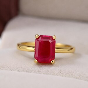 Puede incluir: Un anillo de oro con una gran piedra preciosa roja de corte esmeralda rectangular, sujeta por cuatro pequeñas garras de oro. La banda del anillo es de oro liso y pulido, creando un diseño sencillo que resalta la vibrante piedra roja.